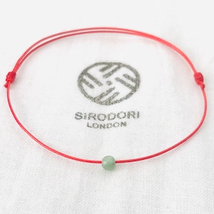 May include: A red string bracelet with a single green jade bead. The bracelet is tied with a knot at each end.
