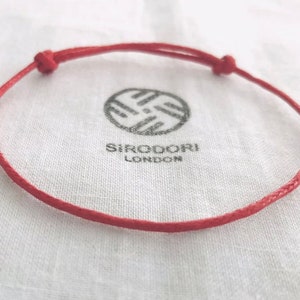 May include: A red adjustable bracelet with a knotted closure. The bracelet is made of a thin, woven material. The bracelet rests on a white cloth with the brand name "SIRODORI LONDON" printed in black.
