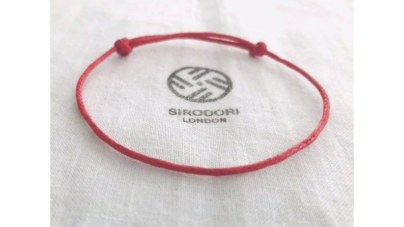May include: A red adjustable bracelet with a knotted closure. The bracelet is made of a thin, woven material. The bracelet rests on a white cloth with the brand name "SIRODORI LONDON" printed in black.
