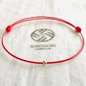 May include: A red string bracelet with a small gold bead. The bracelet is tied with a knot at each end. The bracelet is on a white fabric background. The text "SIRQODORI LONDON" is visible in the background.