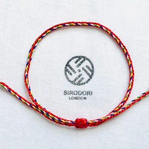 May include: A red, white, blue, yellow and green braided string bracelet with a knot at the end. The bracelet is on a white background.