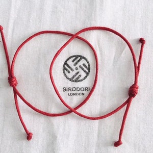 May include: Two red string bracelets tied together with a knot. The bracelets are made of thin cord and have a small bead at each end. The bracelets are on a white background with the text "SIRODORILONDON" printed below.
