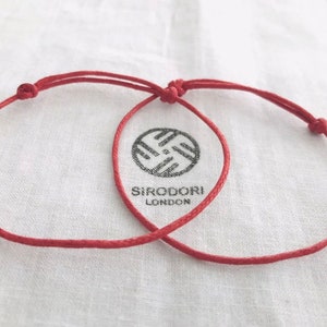 May include: Two red string bracelets with adjustable knots, displayed on a white cloth. The bracelets are intertwined. The cloth has a logo that reads "SIRODORI LONDON".