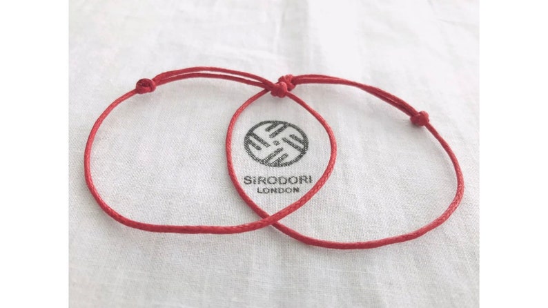 May include: Two red string bracelets with adjustable knots, resting on a white cloth. The bracelets are intertwined, suggesting a connection. The cloth has a logo that reads "SIRODORI LONDON".