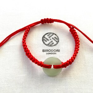 May include: Red string bracelet with a white jade charm. The charm has a hole in the centre. The bracelet is tied with a knot and has two white beads on the ends.