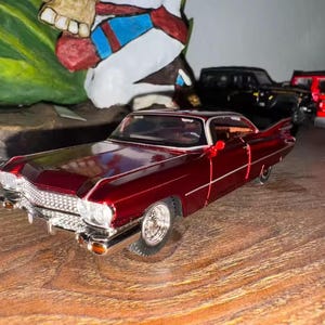 May include: A burgundy classic car model with chrome detailing. The car has a white roof and is set against a blurred background with other toy vehicles. The model car is a miniature replica.