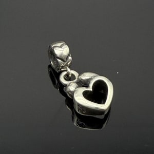 Puede incluir: Un charm plateado con un colgante en forma de corazón. El charm presenta un pequeño diseño detallado en la parte superior, conectado a un corazón más grande con un centro hueco. El metal tiene un acabado pulido, que refleja la luz.