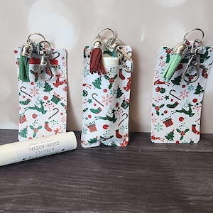 May include: Three Christmas-themed lip balm holders with keychains and tassels. The holders feature a white background with red, green, and brown holiday patterns. A tube of lip balm with text is in front.