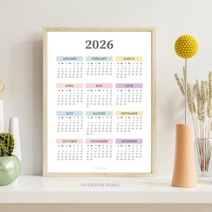 May include: A framed 2026 calendar with pastel-colored month blocks. The calendar is displayed on a white shelf, alongside decorative vases and yellow floral accents. The text "2026" is at the top of the calendar.