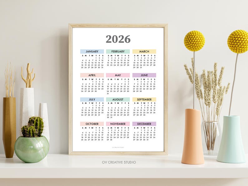 May include: A framed 2026 calendar with pastel-colored month blocks. The calendar is displayed on a white shelf, alongside decorative vases and yellow floral accents. The text "2026" is at the top of the calendar.