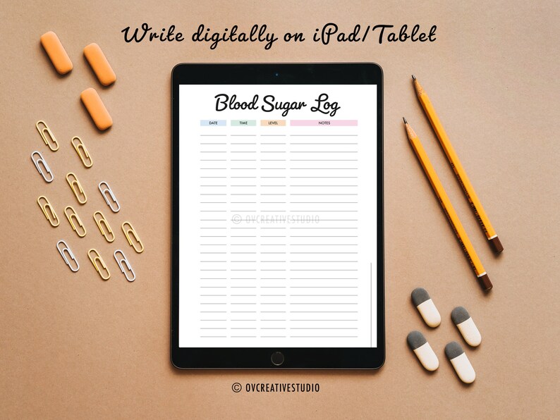 May include: A digital tablet displaying a printable blood sugar log with the text "Blood Sugar Log" in pink and white. The tablet is on a brown surface with yellow pencils, orange erasers, and gold and silver paper clips.