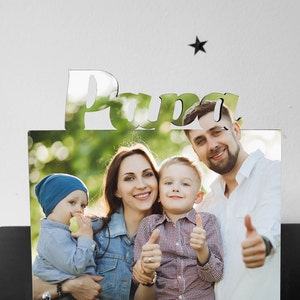 Könnte beinhalten: Ein Fotorahmen mit dem Wort "Papa" ausgeschnitten oben. Der Rahmen enthält ein Foto einer vierköpfigen Familie, darunter ein Vater, eine Mutter und zwei kleine Jungen. Die Familie lächelt und zeigt den Daumen nach oben. Das Foto wurde im Freien in einer grünen Umgebung aufgenommen.