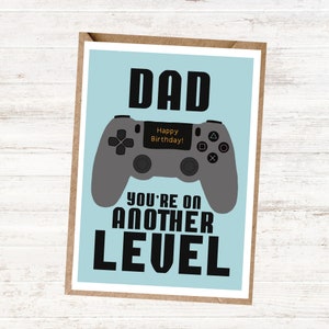 May include: A blue card with a grey video game controller and the text "DAD YOU'RE ON ANOTHER LEVEL" in black. The controller has a screen that says "Happy Birthday!" in orange text.