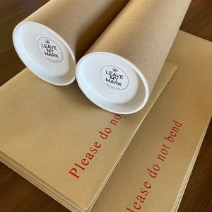 May include: Two brown cardboard tubes with white end caps. The end caps have a white sticker with a black logo that says "Leave My Mark Designs". The tubes are stacked on top of each other. The tubes are on top of two brown cardboard envelopes. The envelopes have red text that says "Please do not bend".