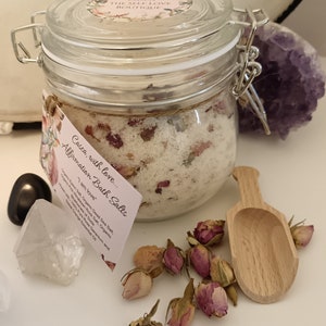 May include: A glass jar filled with white bath salts and dried rose petals. The jar has a metal clasp and is labeled "Casca, with love... Affirmation Bath Salts - "I am love". The label also lists the ingredients: Himalayan Rock Salt, Organic Epsom Salt, Organic Dead Sea Salt, Rose Geranium and Orange Essential Oils.