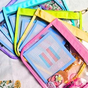 May include: A collection of rectangular clear vinyl pouches with colorful fabric borders in shades of pink, yellow, lime green, and light blue. Each pouch contains a square piece of white needlepoint canvas with a blue and pink striped pattern.