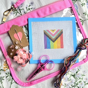 May include: A clear vinyl pouch with pink trim containing a needlepoint canvas featuring a chevron pride flag design, a pair of translucent pink scissors, a bundle of multicolored embroidery floss, and a pink bow on a cardboard tag.
