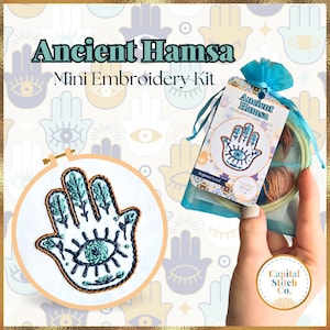 May include: A mini embroidery kit featuring a blue and white Hamsa hand with an evil eye design. The kit includes embroidery floss, a needle, and a small hoop. The kit is packaged in a clear plastic bag with a blue ribbon.