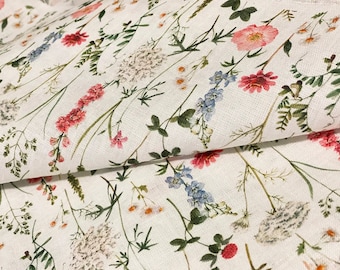 May include: White fabric with a delicate floral pattern featuring pink, red, blue, and white wildflowers and green foliage. The fabric is folded, showing the detailed print of the botanical design.