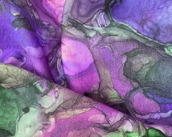 May include: Close-up of a fabric with a watercolor-like design. The fabric features a blend of purple, pink, and green hues, with darker shades creating depth. The material appears to be soft and flowing, with visible texture.