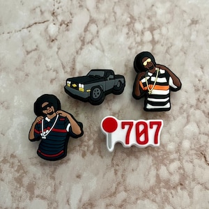 May include: A collection of shoe charms featuring a black pickup truck, two figures with sunglasses and gold chains, and a charm with the number 707 in red and white. The charms are made of a rubber-like material.