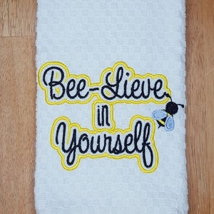 May include: White kitchen towel with yellow and black embroidery that reads "Bee-lieve in yourself" with a small embroidered bee.