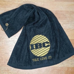 May include: Black towel with gold embroidered "IBC" logo inside a circle with horizontal lines. The text "YULE LOVE IT!" is below the logo. A metal ring is attached to one corner.