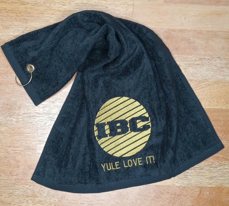 May include: Black towel with gold embroidered "IBC" logo inside a circle with horizontal lines. The text "YULE LOVE IT!" is below the logo. A metal ring is attached to one corner.