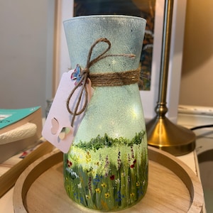 May include: A hand-painted glass vase depicting a meadow. The vase is pale blue at the top, fading to green with colourful flowers at the base. It features a twine bow and a tag with butterfly cutouts. The vase is illuminated with small lights.