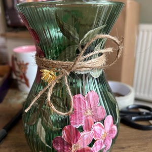 May include: A green ribbed glass vase featuring hand-painted pink flowers and green leaves. The neck of the vase is wrapped with natural jute twine tied in a bow, accented by a small, gold-coloured metal bee charm attached to the twine.