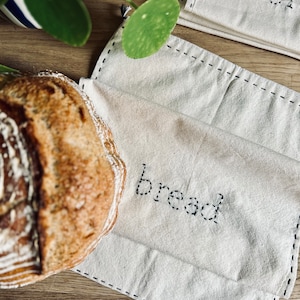 May include: A white cotton bread bag with black stitching and the word "bread" embroidered on it. The bag is lying on a wooden surface next to a loaf of bread.