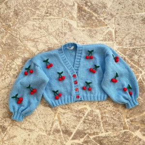 May include: A light blue knitted cardigan with a V-neck and button closure. The cardigan is decorated with red cherries.