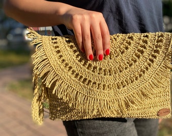 May include: A handmade, light tan crochet clutch with a fringe detail. The clutch features a flap closure and a small leather tag that reads "Handmade". The bag is made of a woven material and has a geometric pattern.