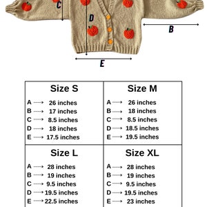 May include: Size chart for a beige knitted cardigan with orange embroidered apples. The chart shows measurements in inches for sizes S, M, L, and XL. Measurements include: A - total length, B - sleeve length, C - shoulder width, D - bust, and E - cardigan length.
