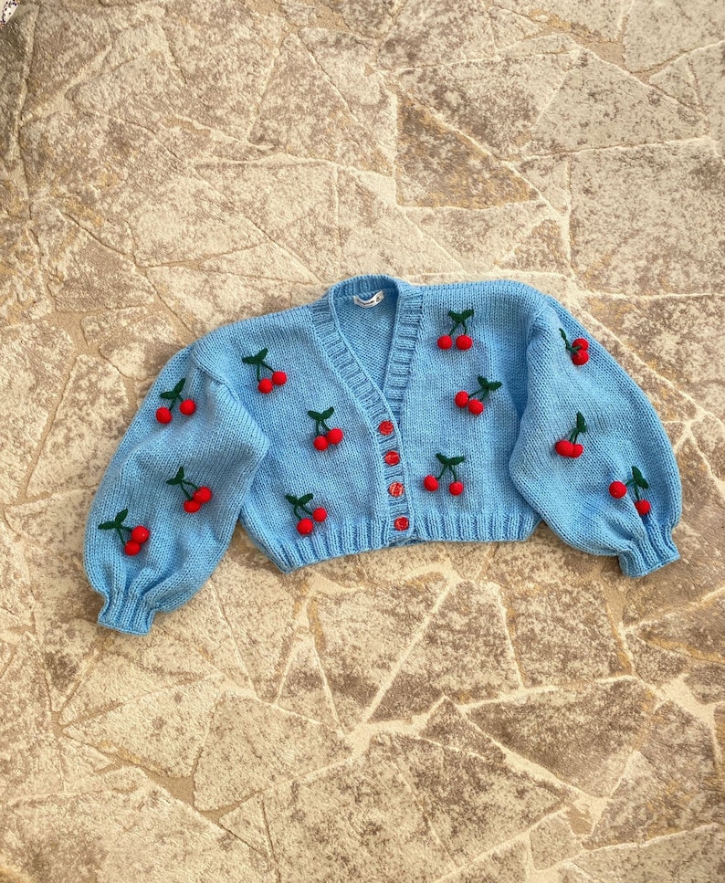 May include: A light blue knitted cardigan with a V-neck and button closure. The cardigan is decorated with red cherries.