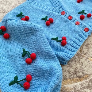May include: A light blue knitted cardigan with a V-neck and button fastening. The cardigan is decorated with embroidered red cherries with green stems.