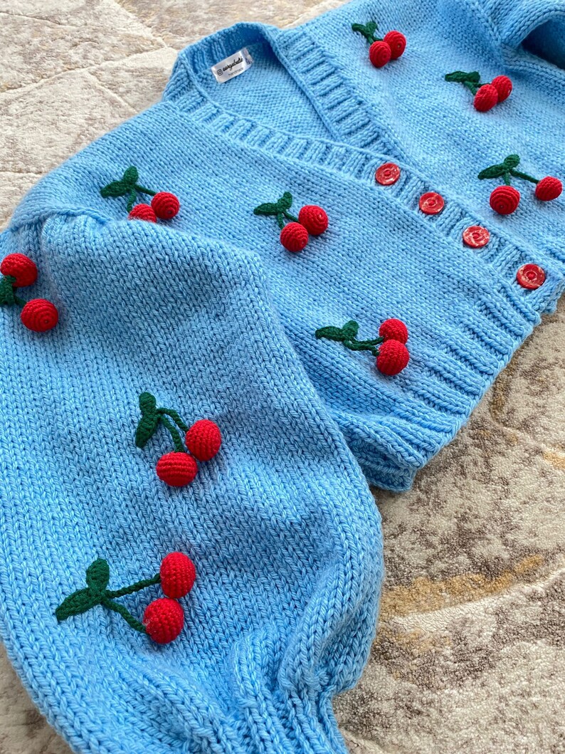 May include: A light blue knitted cardigan with a V-neck and button fastening. The cardigan is decorated with embroidered red cherries with green stems.