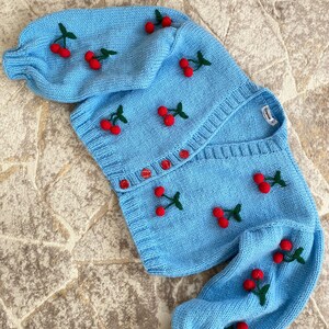 May include: A light blue knitted cardigan with long sleeves and a button-up front. The cardigan is decorated with embroidered red cherries with green stems.
