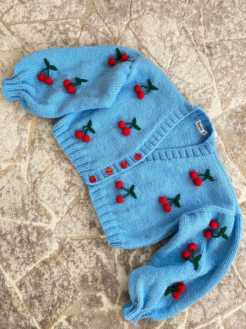 May include: A light blue knitted cardigan with long sleeves and a button-up front. The cardigan is decorated with embroidered red cherries with green stems.