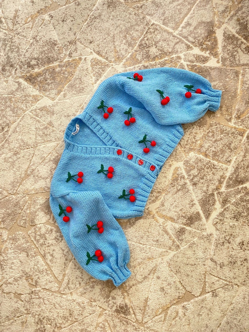 May include: A light blue knitted cardigan with a cherry design. The cardigan has long sleeves and a button closure.