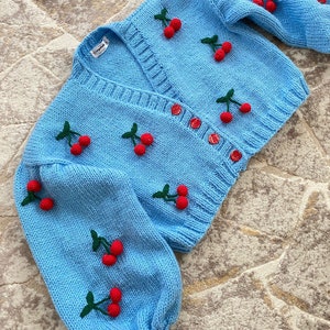 May include: A light blue knitted cardigan with a V-neck and button fastening. The cardigan is decorated with red and green embroidered cherries.