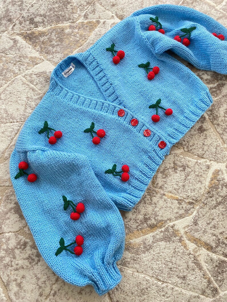 May include: A light blue knitted cardigan with a V-neck and button fastening. The cardigan is decorated with red and green embroidered cherries.