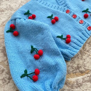 May include: A light blue knitted cardigan with a V-neck and long sleeves. The cardigan is decorated with embroidered red cherries with green stems.