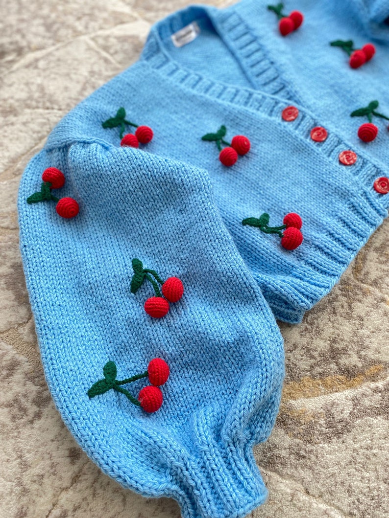May include: A light blue knitted cardigan with a V-neck and long sleeves. The cardigan is decorated with embroidered red cherries with green stems.