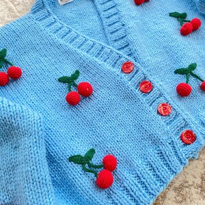 May include: A light blue knitted cardigan with a V-neck and button closure. The cardigan is decorated with embroidered cherries in shades of red and green.