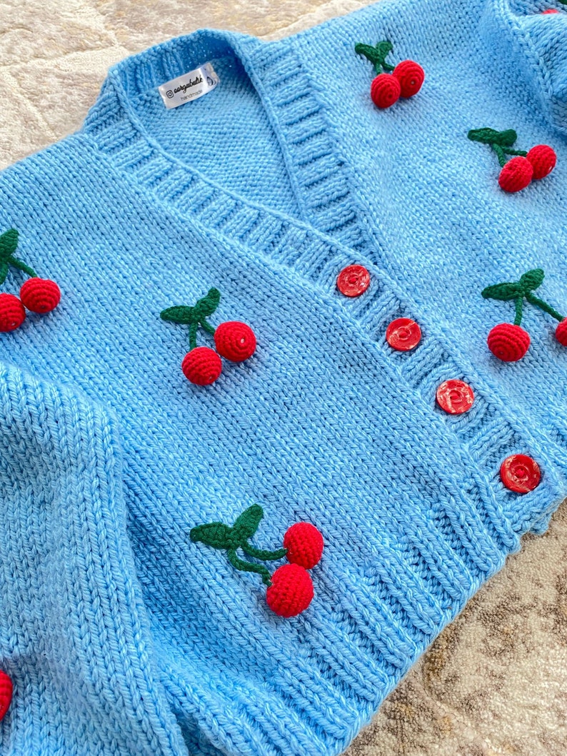 May include: A light blue knitted cardigan with a V-neck and button closure. The cardigan is decorated with embroidered cherries in shades of red and green.