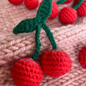 May include: A close-up of two crocheted cherries with green stems and leaves. The cherries are red and have a textured surface. The cherries are on a pink knitted fabric.