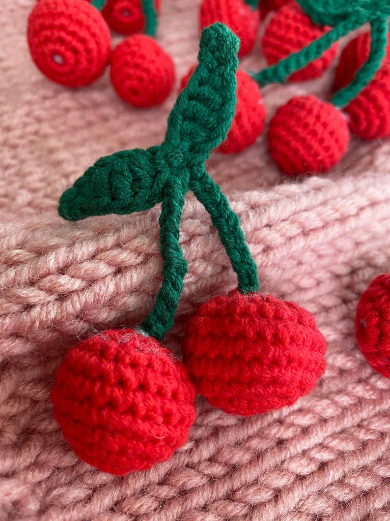 May include: A close-up of two crocheted cherries with green stems and leaves. The cherries are red and have a textured surface. The cherries are on a pink knitted fabric.