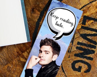 May include: A blue and white bookmark with a watercolor background. A man with short brown hair is featured on the bookmark. A speech bubble with the text "Keep reading, babe." is above the man's head.
