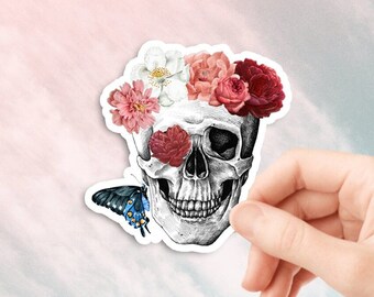 May include: A white skull with a floral crown of pink, red, and white flowers. A blue butterfly is perched on the skull's jaw.